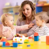 Nauczycielka i dwoje dzieci bawią się klockami przy żółtym stole w żłobku.